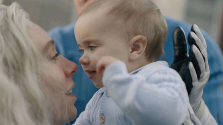 A imagem mostra Sue e um Franklin, próximos um do outro, em um momento de afeição e carinho. A composição da imagem é centrada no encontro próximo entre a mulher e o bebê. A mulher está olhando para o bebê, seus rostos estão próximos. O bebê está sendo segurado pela mulher. O foco está principalmente nos rostos da mulher e do bebê, enquanto o fundo é desfocado. A imagem sugere intimidade e carinho. Seu rosto e expressões demonstram emoção e carinho. Franklin é um bebê, de pele clara e cabelos claros. Seu rosto é incrivelmente expressivo e mostra um sorriso suave e genuíno, expressando alegria. A luz natural cria um ambiente suave e aconchegante, focando na conexão entre as duas pessoas. A atmosfera é de ternura e afeto materno.