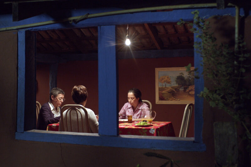 Imagem do filme Nada. A cena é observada do exterior de uma casa, através de uma janela retangular dividida por duas colunas de madeira pintadas de azul vivo. No interior, três mulheres estão reunidas em torno de uma mesa coberta por uma toalha estampada com motivos florais vermelhos e verdes. Uma lâmpada incandescente pendurada no teto ilumina a cena. À esquerda, uma mulher com cabelos curtos e óculos veste um casaco vinho sobre uma blusa clara. Ao centro, de costas para o observador, outra mulher com cabelos castanhos curtos veste um suéter branco texturizado. À direita, uma terceira mulher com cabelos escuros presos veste uma camisa listrada em tons de rosa e branco e parece estar escrevendo ou lendo algo sobre a mesa. Sobre a mesa, notam-se objetos como copos, jarras e um recipiente com alimento.