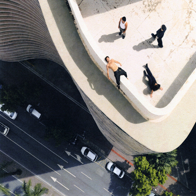 A capa do álbum é uma fotografia aérea de um prédio com design moderno e curvas suaves, vista de cima. Quatro homens estão sobre a borda do edifício, em poses descontraídas: um está deitado com o peito nu, outro está sentado no chão com as pernas esticadas, e dois caminham pelo espaço. Abaixo, é possível ver a rua com carros brancos e árvores margeando a calçada. A cena sugere um ensaio fotográfico ousado, com forte contraste entre o ambiente urbano e a sensação de liberdade dos modelos.