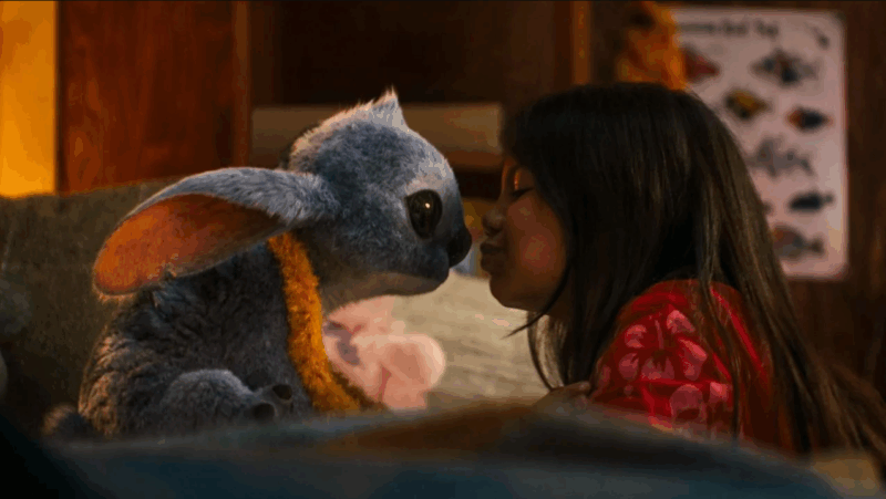 Cena do filme live-action mostrando Lilo, uma menina de cabelos longos, vestindo uma camisa vermelha com estampas florais brancas, inclinando-se para dar um beijo no nariz de Stitch, o personagem alienígena azul. Stitch tem uma aparência felpuda e realista, com orelhas grandes e olhos expressivos. Ele está usando um colar de flores amarelas e ambos estão em um quarto com um pôster embaçado ao fundo. A cena transmite um momento de carinho e conexão entre os dois personagens.