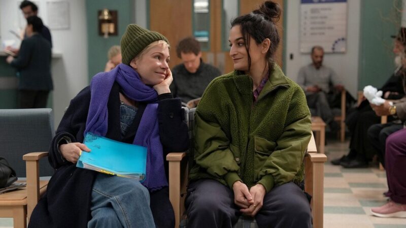 Cena da série Morrendo por Sexo. Molly, loira e de cabelos curtos veste um cachecol roxo e um gorro verde escuro e segura uma pasta azul enquanto olha para sua amiga. Na direita, Nikki, vestindo um casaco verde sorri para Molly. As duas estão sentadas em uma sala de espera de um hospital.