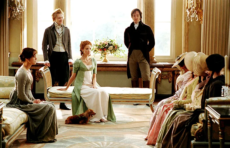 Cena do filme Orgulho e Preconceito (2005). Na imagem, vemos uma elegante sala de estar em tons dourados e claros, com vários personagens vestidos com roupas de época. Ao centro, uma mulher ruiva está sentada com um vestido verde e branco, com um pequeno cachorro marrom deitado aos seus pés. Atrás dela, dois homens em pé vestem roupas de época — casacos compridos e calças altas. À esquerda, uma mulher sentada observa a cena, enquanto à direita, um grupo de mulheres com vestidos coloridos e chapéus senta-se em um sofá comprido. A cena transmite formalidade e tensão social