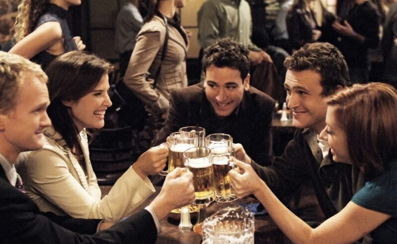 Frame de cinco amigos, Barney, homem loiro vestindo terno e gravata, Robin, mulher morena de jaqueta bege, Ted, homem branco de cabelo preto usando um blazer marrom, Marshall, homem branco com entradas no couro cabeludo e uma camiseta marrom, e Lily, mulher ruiva usando uma blusa azul, em uma mesa de bar sorrindo e brindando com canecas de cerveja. No fundo, figurantes circulando pelo ambiente.