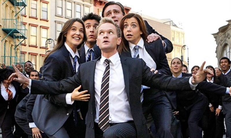 Frame dos cinco amigos, Barney, Ted, Marshall e Lily,da esquerda para a direita, trajados em terno e gravata, enquanto Barney, homem branco e loiro, se posiciona à frente deles com os braços abertos. Ao fundo, vários figurantes vestidos da mesma forma preenchem o quadro.