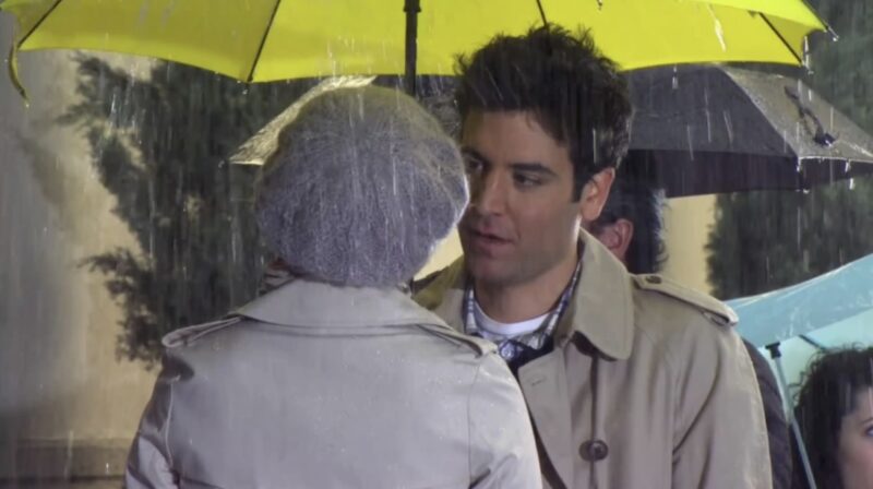 Frame de Ted Mosby, homem branco de cabelos escuros, vestindo um sobretudo bege, enquanto segura um guarda-chuva amarelo, conversando com uma mulher que está de costas e usa também um sobretudo bege junto de um gorro cinza.