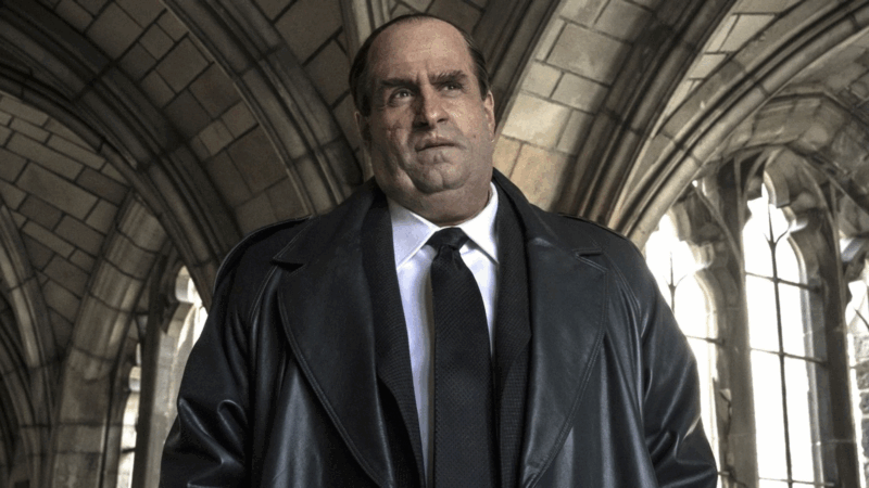 Cena da série Pinguim. A imagem mostra Oswald Cobblepot de frente, vestindo um casaco de couro preto, camisa branca e gravata escura, em um ambiente de arquitetura gótica com arcos de pedra e janelas altas. Ele encara algo fora de cena com uma expressão rígida e desconfiada, transmitindo tensão e autoridade. A luz suave que entra pelas janelas destaca o contraste entre a frieza do cenário e o tom sombrio do personagem, reforçando o clima dramático da narrativa.