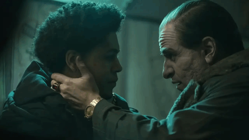 Cena da série Pinguim. Em um ambiente escuro iluminado por luz esverdeada, Oswald Cobblepot segura o rosto de uma jovem com ambas as mãos, olhando fixamente em seus olhos. Ele usa um casaco grosso com gola de pele e está com cabelo penteado para trás. Em suas mãos, ele exibe um anel dourado e um relógio de pulso da mesma tonalidade. A jovem, de pele escura e cabelo crespo, encara de volta com seriedade. A cena transmite tensão e emoção contida, sugerindo uma conversa íntima ou momento de confronto.