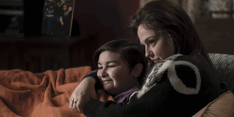 Cena da série Pinguim. Uma mulher e um menino estão sentados juntos em um sofá, cobertos por uma manta laranja. A mulher envolve o menino com os braços, enquanto ele sorri levemente, demonstrando conforto e afeto. Ambos observam algo fora de cena. Ao fundo, em uma prateleira escura, há uma fotografia emoldurada com a imagem de três meninos. A cena transmite um momento íntimo de conexão familiar, segurança e ternura.