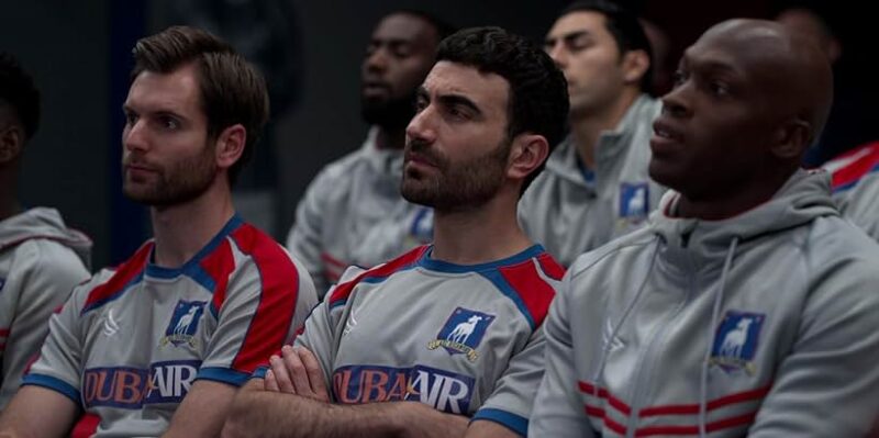 Frame de Roy Kent, homem branco, de cabelo e sobrancelha preta, barba e bigode por fazer, vestindo a camisa de treino com o escudo azul do time do AFC Richmond cinza com mangas em detalhes vermelhos e linhas azuis junto de um patrocínio da Dubai Air – marca fictícia –, em meio a seus dois companheiros de equipe, à esquerda um homem branco de cabelo, bigode e barba castanhos e à direita um homem negro de cabelo raspado, com a mesma tonalidade de roupa.