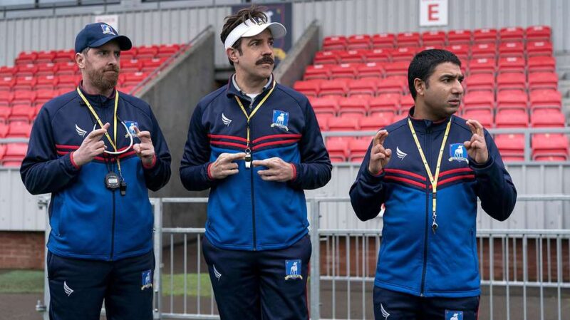 Frame de Ted Lasso, homem branco de cabelo liso castanho tapado pela viseira branca e bigode alinhado, em meio a seus companheiros Beard, homem branco de cabelo e barba ruivos, e Nate, homem baixo e moreno, todos vestindo um uniforme azul marinho e celeste com listras vermelhas. Ao fundo, uma arquibancada cinza com cadeiras vermelhas em um centro de treinamento com grama. 