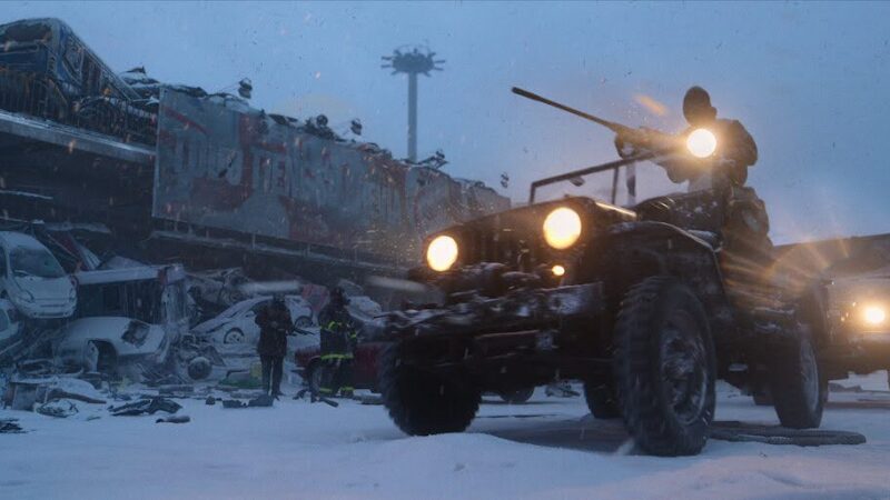 cena da série O Eternauta. A cena mostra em primeiro plano um jipe das forças armadas argentinas com um soldado posicionado acima do veículo, portando um rifle longo. Ao fundo, a parte inferior de um viaduto está fechado com uma pilha de carros destruídos formando uma barricada. A rua está coberta de neve e a paisagem está envolta em névoa, com um céu acinzentado
