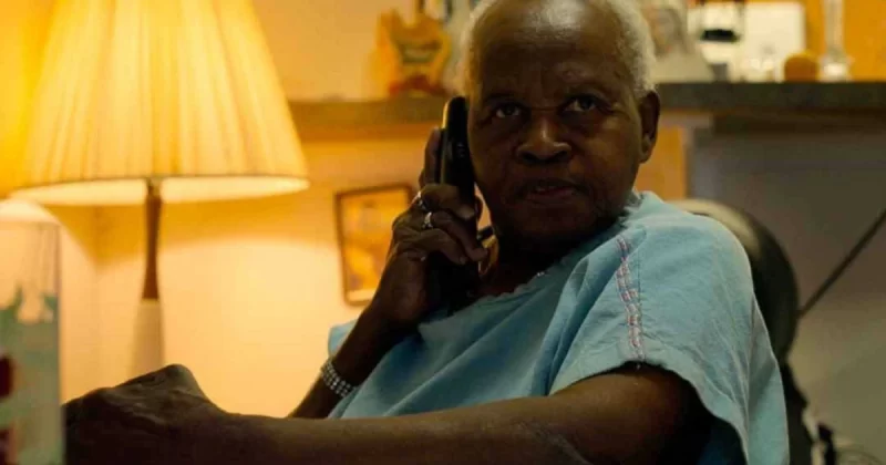 Cena do documentário A Mulher da Casa Abandonada. Fotografia de Hilda dos Santos, uma mulher negra de cabelo curto e branco, vestindo uma blusa azul, segurando um telefone sem fio próximo ao ouvido. No fundo, aparece um abajur ligado e alguns itens de decoração.