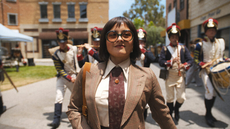 Cena da série O EstúdioNa imagem, a atriz Chase Sui Wonders está andando, com um sorriso de canto no rosto, esboçando confiança. Atrás dela, há homens vestindo uniformes de militares de séculos passados. Ela é uma mulher na faixa dos 30 anos, está usando um óculos redondo de sol, com lente alaranjada, terno com linhas na cor marrom e gravata na mesma cor, com uma estampa de olhos. Seu cabelo é preto, curto e com franja. O dia está ensolarado. 