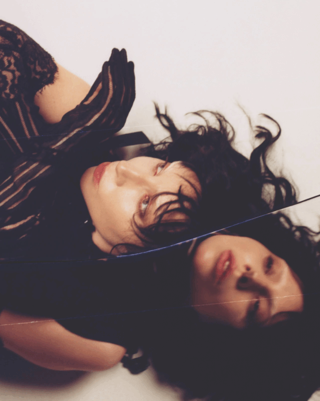 Lady Gaga aparece deitada de lado, com os cabelos soltos e escuros espalhados ao redor, usando uma roupa preta rendada. Seu rosto se reflete em uma superfície espelhada abaixo dela, criando um efeito duplicado e onírico. A fotografia, com atmosfera etérea e melancólica, integra a série de imagens de divulgação de MAYHEM (2025).