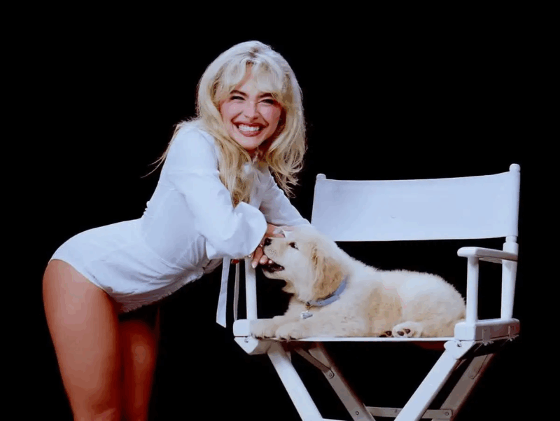 A imagem mostra Sabrina Carpenter e um filhote canino de Golden Retriever, retratados em close-up contra um fundo preto, com foco total na interação entre eles. A mulher, sorridente e vestida de branco, aparece inclinada à esquerda, enquanto o cachorro está relaxado sobre uma cadeira de diretor branca no centro da cena. A iluminação suave e sem sombras fortes destaca a expressão alegre da mulher e o ar descontraído do animal, transmitindo uma atmosfera de carinho, ternura e sofisticação.