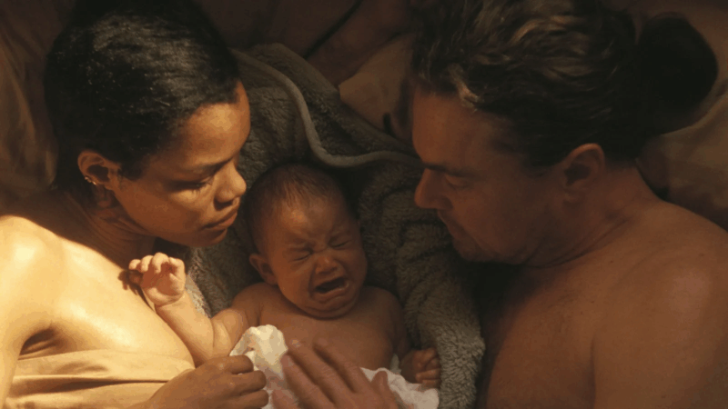 Cena do filme Uma Batalha Após a OutraNa imagem, da esquerda para a direita, estão Perfidia, Willa e Bob, eles são uma família. Os três estão deitados em uma cama, sem roupas, Willa, um bebê, está no meio de Bob e Perfidia, chorando. Ela está deitada em uma coberta grossa. Os pais estão com a mão apoiada no corpo dela, em sinal de calma. Raios de sol atingem o peito e ombros de Perfidia, iluminando a cena. Perfidia é uma mulher negra na faixa dos 35 anos, usa cabelo curto. Bob é um homem branco, na faixa dos 50 anos, de cabelos lisos e claros. 