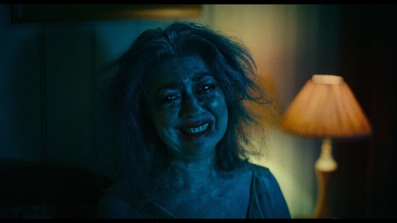 Cena do filme JuntosNa imagem, uma senhora está sentada em uma cama enquanto chora. Ela está com um sorriso no rosto e batom borrado nos lábios. Seus cabelos, grisalhos, estão bagunçados. Ao seu lado direito, há um abajur de luz amarela ligado. Seu rosto é iluminado por um azul noturno. 