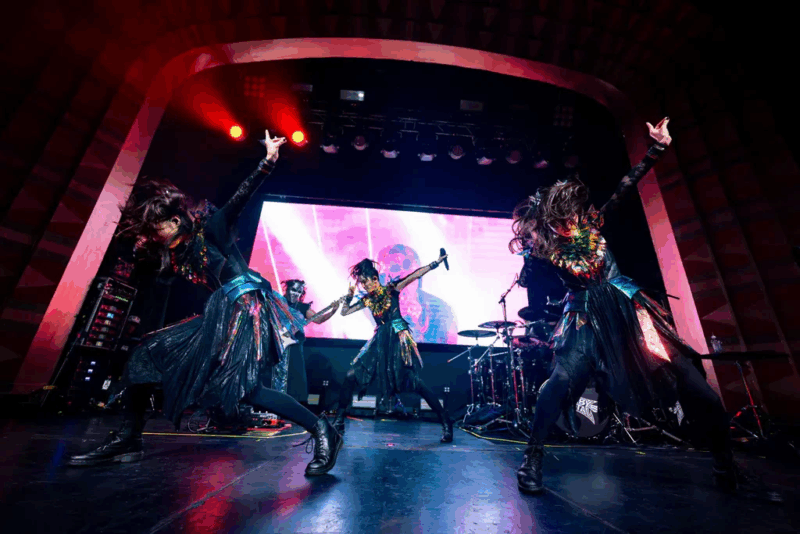 Na imagem de um show da banda, as três integrantes Momometal, Su-metal e Moametal, respectivamente, se apresentam em um palco iluminado com luzes vermelhas e um telão colorido ao fundo. Elas usam trajes pretos com detalhes cintilantes e fazem coreografias energéticas, com braços erguidos e cabelos em movimento, transmitindo intensidade.
