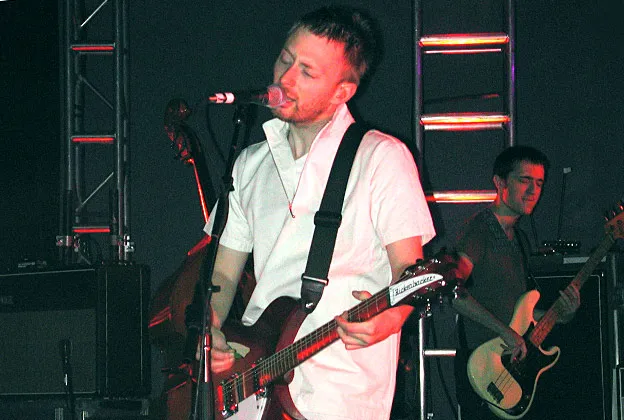 Um cantor toca guitarra elétrica enquanto canta em um microfone durante um show. Ele usa uma camisa branca de manga curta e segura uma guitarra vermelha. A iluminação do palco é escura, com luzes verticais coloridas ao fundo. Ao fundo, um baixista também toca seu instrumento.