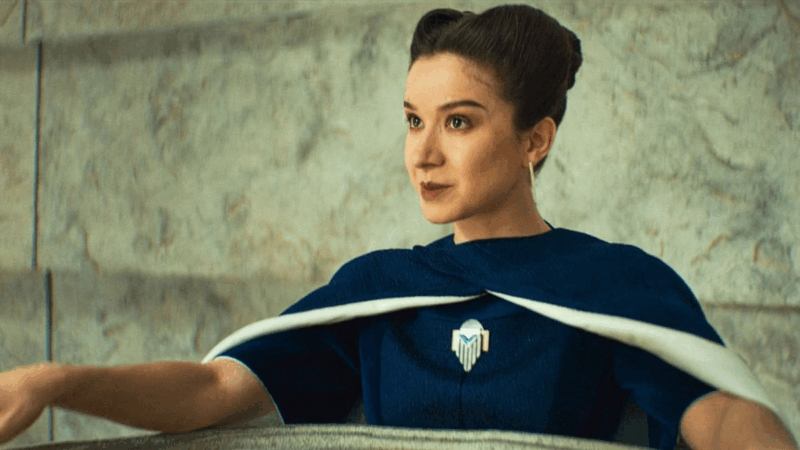 No centro da imagem está Kleya, uma mulher branca de cabelos pretos e penteados para trás. A personagem está maquiada e possui olhos castanhos. Ela utiliza um vestido azul com um broche geométrico no centro do peito e uma capa azul e branca. Ela conversa com um personagem fora do plano e sorri. Atrás dela há uma parede branca com aspecto de mármore.