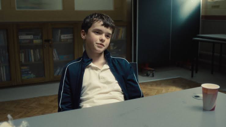 Cena da série Adolescência. Jamie, garoto de cabelo escuro e curto, está sentado em uma mesa no centro da imagem, levemente inclinado para trás, com uma expressão desafiadora no rosto. Ele veste uma jaqueta azul-escura com listras brancas nas mangas e uma camisa polo branca por baixo. Ao fundo, há um armário com livros. A luz entra parcialmente pela lateral. Em cima da mesa, à direita, há um copo descartável.
