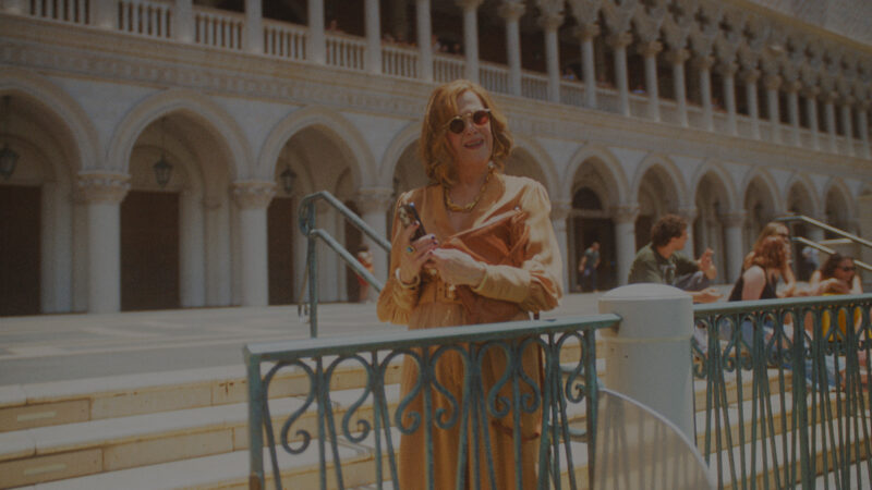 Cena da série O EstúdioNa imagem, a atriz Catherine O'Hara está em pé, parada em frente a uma grade que separa chão e rio. Ela segura um celular na mão e está vestindo um vestido laranja. Nos braços, ela também carrega uma bolsa na mesma cor, além de usar óculos escuros em formato redondo no rosto. Ela é uma mulher branca, na faixa dos 70 anos, com cabelos ruivos. Ao fundo, há uma escada, onde jovens estão sentados e conversando, no canto direito da imagem. 
