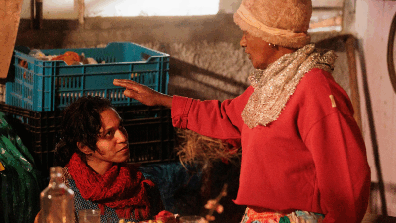 Na imagem, Dora, uma mulher de pele escura e cabelos cacheados, está sentada e olha para uma mulher mais velha que está em pé à sua frente. Dora veste um casaco azul estampado e um cachecol vermelho vibrante. A mulher mais velha, também de pele escura, usa um gorro macio, um suéter vermelho folgado e um cachecol brilhante, e tem a mão direita estendida sobre a cabeça de Dora, em um gesto que parece de bênção ou acolhimento. Ao fundo, caixas plásticas azuis e verdes sugerem um ambiente doméstico ou de armazenamento.