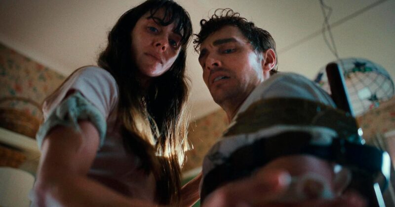 Cena do filme JuntosNa imagem, com a visão de baixo para cima, Millie e Tim olham com expressão de medo para seus braços colados. À direita, está Tim anexado com fita adesiva em uma cadeira de cozinha. Millie está à esquerda, no colo do namorado. Ambos vestem camiseta branca e possuem pele clara. Tim é um homem na faixa dos 40 anos, de cabelos claros e corte que parece um mullet. Já Millie é uma mulher na faixa dos 40 anos, de cabelos escuros e longos. Ela usa franja na testa. Os dois personagens estão suados e descabelados. 