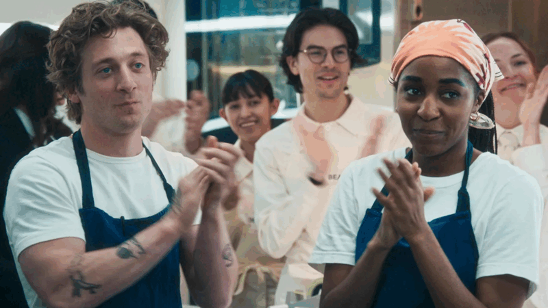 Em uma cena da série The Bear, Carmy (à esquerda) e Sydney (à direita) estão em primeiro plano, ambos sorrindo e batendo palmas. Carmy usa uma camiseta branca e um avental azul. Sydney usa uma bandana laranja, camiseta branca e um avental azul combinando. Ao fundo, outros membros da equipe da cozinha também estão sorrindo e aplaudindo, criando uma atmosfera de celebração e sucesso compartilhado.