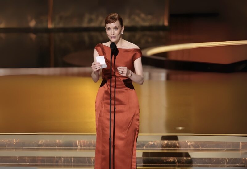 Britt Lower vestindo um elegante vestido longo de cor terracota com detalhes assimétricos. Ela está segurando um pequeno cartão ou papel na mão direita e falando em um microfone em um pedestal. O fundo é um cenário dourado e reflexivo. A iluminação destaca o vestido e cria um efeito brilhante no ambiente ao redor.