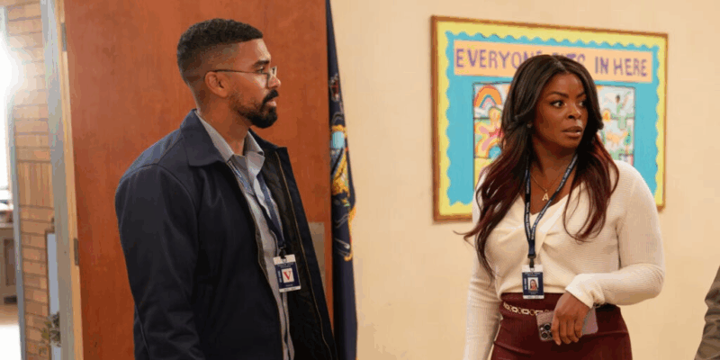 Na imagem, da esquerda para a direita, há um homem negro de cabelo curto usando óculos e vestindo um casaco azul-marinho e uma camiseta social cinza, com um crachá no pescoço. Ao seu lado, há uma mulher negra de cabelo longo e liso, usando um crachá no pescoço, uma blusa de manga longa branca, uma saia vermelha-escura e um cinto dourado. Em sua mão esquerda, há um celular. 