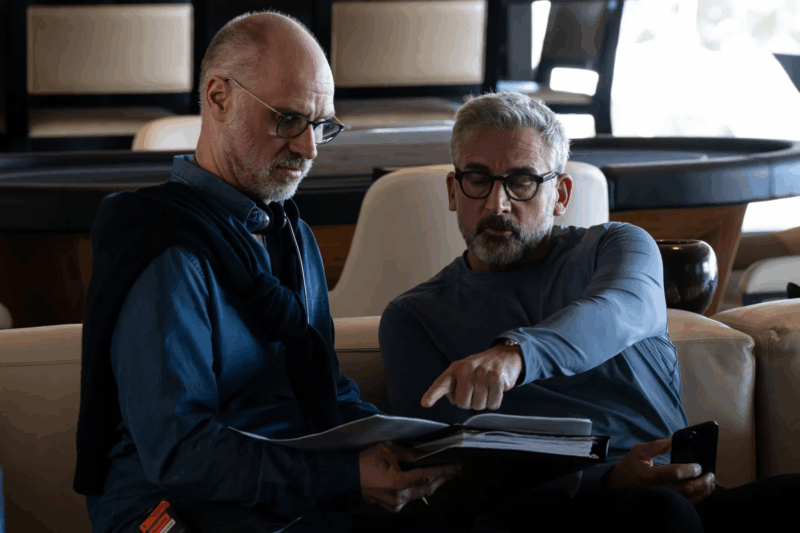Na foto, dois homens estão sentados em um sofá. Um segura uma pasta aberta, enquanto o outro aponta para algo entre as páginas. Ambos usam óculos e expressam concentração, como se analisassem documentos importantes em um ambiente de reunião.