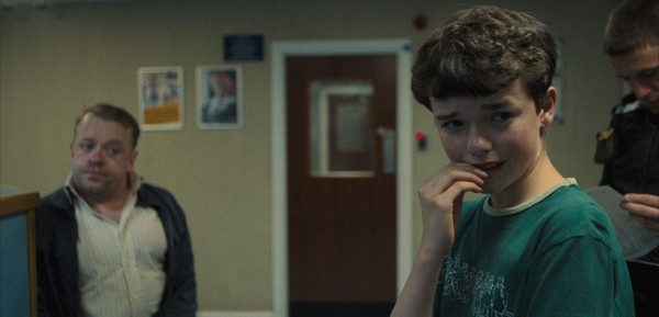 Cena da série Adolescência. No canto direito, vemos Jamie, de cabelo escuro e curto, vestindo uma camiseta verde. Ele olha para o lado com uma expressão preocupada e a mão próxima à boca, em um gesto de nervosismo. No centro, vemos uma porta de madeira, com uma janela, na parede ao lado dela, dois quadros. Atrás de Jamie, quase fora do quadro, aparece um policial uniformizado olhando para um bloco de anotações. À esquerda, há um homem de meia-idade com expressão séria, vestindo uma camisa branca e jaqueta azul-escura.