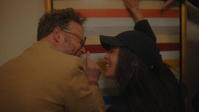  Cena da série O EstúdioNa imagem, os atores Seth Rogen e Zoë Kravitz estão rindo enquanto tocam em um quadro de faixas coloridas. Ele é um homem branco na faixa dos 40 anos, de cabelos curtos e grisalhos. Está vestindo um terno amarelo. Já a atriz, é uma mulher negra, na faixa dos 35 anos, de cabelos escuros e com tranças longas. Ela usa um boné preto. 