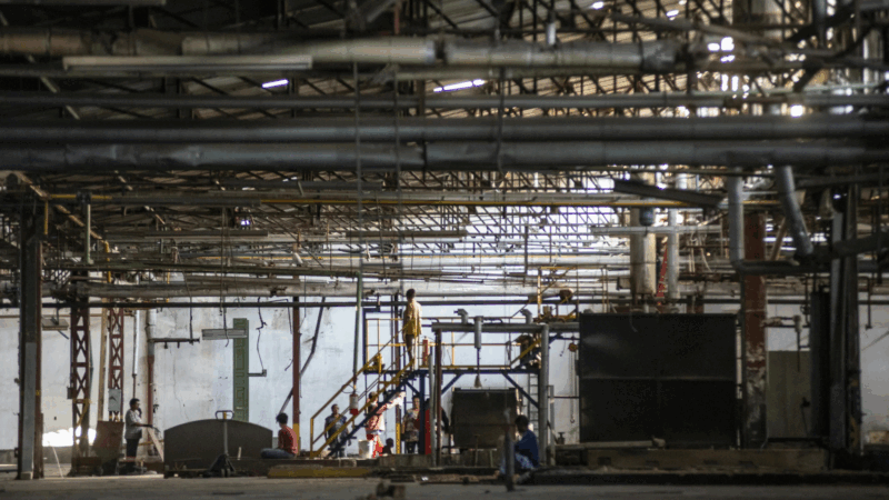 O interior de uma fábrica, com o teto repleto de uma complexa rede de tubulações, vigas e dutos metálicos. A luz entra por algumas aberturas superiores, iluminando parcialmente o ambiente desgastado. Figuras humanas, pequenas em comparação com a imensidão do local, estão espalhadas pelo chão e em uma escadaria de metal no centro da cena.