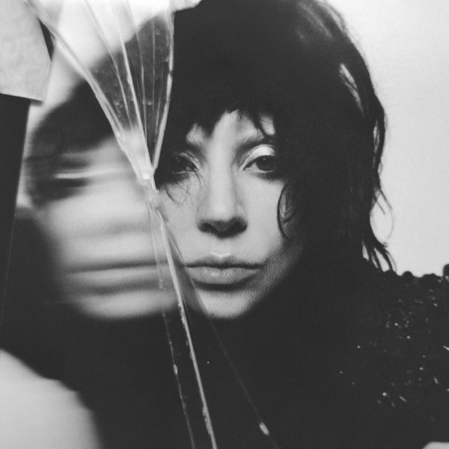 Capa do álbum MAYHEM (2025) de Lady Gaga. Imagem em preto e branco, o rosto da cantora está parcialmente visível através de um vidro quebrado, criando um efeito de distorção e duplicação de sua imagem. Ela tem uma expressão séria, com maquiagem suave e cabelo curto e escuro, bagunçado, caindo sobre os ombros. A composição transmite uma atmosfera introspectiva, artística e levemente melancólica.