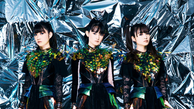 Na imagem, de ensaio fotográfico, as três integrantes Momometal, Su-metal e Moametal, respectivamente, da banda BABYMETAL, posam diante de um fundo metálico prateado. Elas vestem figurinos pretos com detalhes brilhantes em verde, azul e dourado, lembrando escamas. Todas têm expressões sérias, penteados presos e maquiagem discreta.