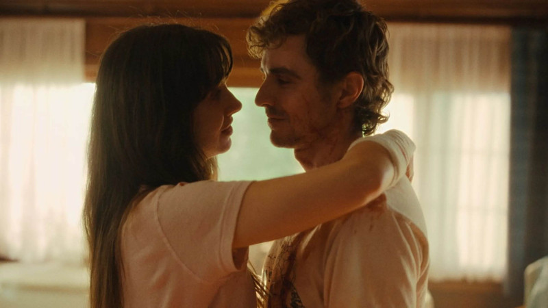 Cena do filme JuntosNa imagem, os personagens Tim e Millie estão de pé, abraçados. Ela, à esquerda, está com o braço direito apoiado atrás do pescoço dele, parte do punho está enfaixado. Ambos os rostos estão próximos. Enquanto ela fala, Tim a encara com ternura. Ambos vestem camiseta branca e possuem pele clara. Tim é um homem na faixa dos 40 anos, de cabelos claros e corte que parece um mullet. Sua roupa e rosto estão manchados de sangue. Já Millie é uma mulher na faixa dos 40 anos, de cabelos escuros e longos. Ela usa franja na testa. 