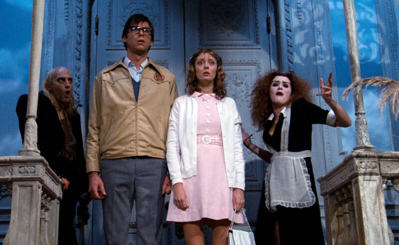Cena do filme The Rocky Horror Picture Show.Na imagem, da esquerda para a direita: Riff Raff, pálido enquanto canta, está parado na entrada do salão principal do castelo, vestindo um terno preto aberto que revela seu peito, calça escura e luvas desgastadas. Ao seu lado, Brad usa uma jaqueta de couro bege sobre um suéter azul claro e camisa branca, calça cinza e óculos. Janet, ao centro, veste um vestido rosa com gola, coberto por um cardigã branco, e segura uma bolsa clara. À direita, Magenta aparece com cabelos desgrenhados e expressão teatral, trajando uma clássica roupa de empregada dos anos 1950 (vestido preto com avental branco rendado). Brad e Janet estão paralisados, olhando para frente com espanto. 