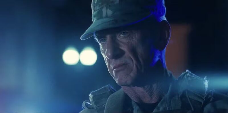 Cena do filme Uma Batalha Após a OutraNa imagem, o personagem Lockjaw está centralizado, encarando à frente com uma expressão de desprezo. Está de noite e a iluminação é azul escuro. Lockjaw é um homem na faixa dos 65 anos, de pele e olhos claros. Veste uniforme e boné do exército. 