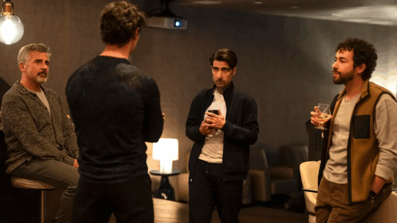 Numa cena do filme, quatro homens conversam em um ambiente interno de iluminação suave. Um deles está sentado em um banco, enquanto os outros permanecem de pé, segurando celular e taças de bebida. As roupas casuais e o cenário intimista sugerem um encontro descontraído.