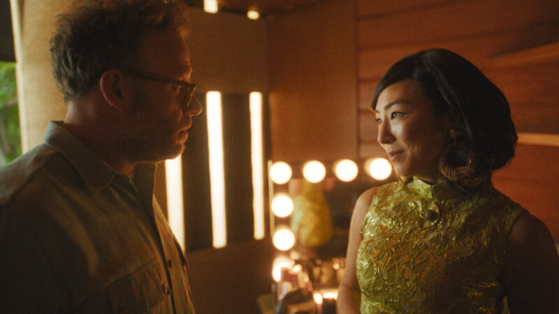 Cena da série O EstúdioNa imagem, os atores Seth Rogen (à esquerda) e Greta Lee (à direita) estão conversando em um set de filmagens. Ela olha com ternura para Rogen, enquanto ele presta bastante atenção nela. Greta Lee é uma mulher com traços asiáticos, de cabelos escuros na altura do pescoço. Usa um vestido verde com detalhes em amarelo e gola alta. Seth Rogen é um homem branco, de cabelos curtos e grisalhos. Ele veste uma camisa de botões e óculos. Atrás dos atores está um espelho com lâmpadas acesas na borda, trazendo uma iluminação quente ao ambiente. 