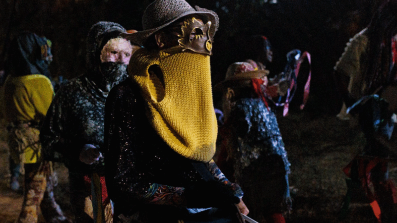 Em uma cerimônia noturna da Guarda de Moçambique Nossa Senhora do Rosário e Santa Efigênia, um participante em primeiro plano está vestido com trajes rituais. Ele usa um chapéu, uma máscara dourada que cobre a área dos olhos e um tecido de lã amarelo que esconde a parte inferior do rosto. Ao seu redor, outras pessoas mascaradas e com vestimentas tradicionais participam da celebração em um ambiente com pouca luz, criando uma atmosfera mística.