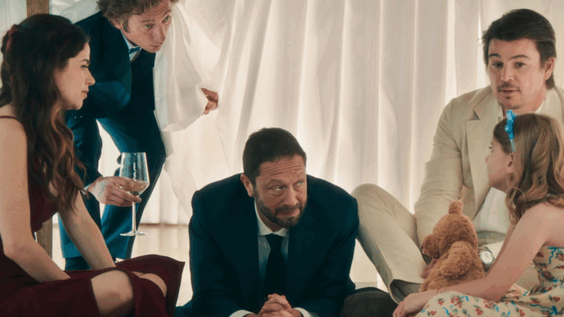 Em uma cena da série The Bear, um grupo de personagens está reunido debaixo de uma mesa. Da esquerda para a direita: a personagem Claire, com um vestido vermelho, está sentada e segura uma taça de vinho. Atrás dela, Carmy, vestindo um terno, inclina-se com uma expressão preocupada. No centro, sentado no chão, está Richie, de terno escuro, conversando com intensidade. Ao seu lado direito está Frank, de terno claro, e na extrema direita, a menina Eva, que segura um ursinho de pelúcia, sentada no chão e interagindo com os outros.