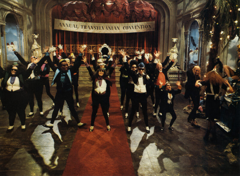Cena do filme The Rocky Horror Picture ShowNa imagem, está o salão principal do castelo durante o número musical "Time Warp". Um grupo de personagens excêntricos, vestidos com smokings coloridos, cartolas e calçados de sapateado, estão com os braços erguidos em perfeita sincronia sobre um tapete vermelho central. Ao fundo, uma faixa pendurada no palco anuncia “Annual Transylvanian Convention” (“Convenção Anual da Transilvânia” em português). As cortinas vermelhas e a arquitetura do salão criam um clima teatral e decadente. À direita, Riff Raff e Magenta dançam juntos. 