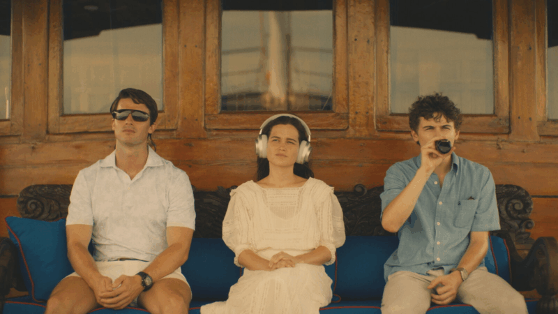 Cena da série The White Lotus. Nela, observa-se três pessoas sentadas em um barco. Da esquerda para a direita, há um homem branco de óculos de sol que utiliza uma camisa e short brancos, uma mulher branca com cabelos castanhos que usa um vestido branco e utiliza um fone de ouvido branco e um homem branco que está tomando uma bebida e veste uma camisa azul e uma calça bege. Os três olham para frente de maneira séria.