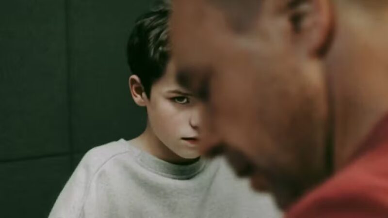 Cena da série Adolescência. Duas personagens aparecem em destaque, o fundo é escuro e neutro, como um ambiente fechado. Na frente, um pouco desfocado, há um homem adulto de perfil, usando uma camisa vermelha, o detetive Luke Bascombe. Seu rosto está parcialmente cortado pela borda direita da imagem. Atrás, de forma mais nítida, está a personagem Jamie Miller, um garoto com expressão séria e olhar fixo, olhando para frente, mas de cabeça baixa. Ele tem cabelo escuro e curto, e veste uma blusa cinza clara. 