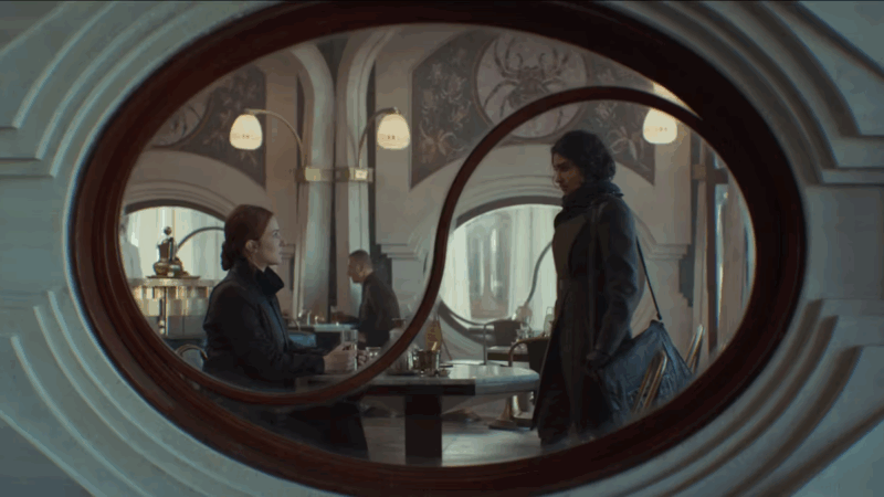 A imagem é enquadrada por uma estrutura oval decorativa. Uma mulher branca de cabelos ruivos e presos e roupa escura está sentada à mesa, à esquerda. É a Vel. Ela está olhando para uma jovem de pele escura de cabelo cacheado preto e roupa escura com uma bolsa lateral, que está de pé. As duas estão enquadradas pela estrutura oval da janela remete ao símbolo de Ying Yang. Ao fundo, fora de foco, está um café.