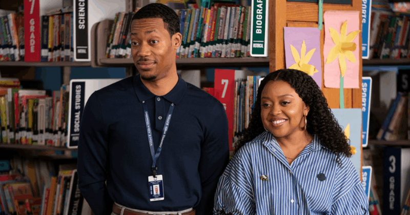 Na imagem, da esquerda para a direita, há um homem negro de cabelo curto sorrindo, usando um suéter azul marinho e um crachá no pescoço. Ao seu lado, há uma mulher negra de cabelo longo e cacheado sorrindo, usando uma blusa listrada de branco e azul e brincos dourados.