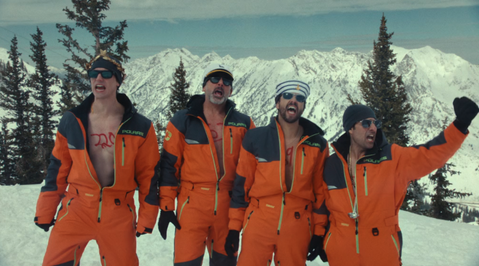 Numa cena do filme, quatro homens vestindo macacões laranja de esqui estão em uma montanha coberta de neve. Alguns estão com os zíperes abertos, exibindo roupas por baixo. Atrás deles, picos nevados completam a paisagem. O grupo transmite energia e empolgação.